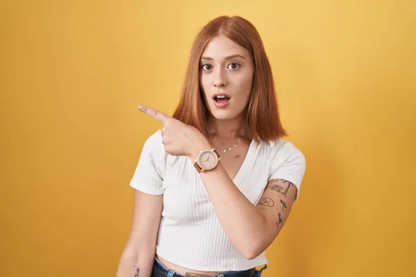 Young Redhead Woman Standing Yellow Background Surprised Pointing Finger Side — Foto de Stock