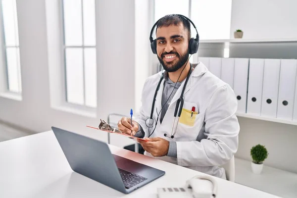 Young hispanic man doctor listening to music writing medical report at clinic