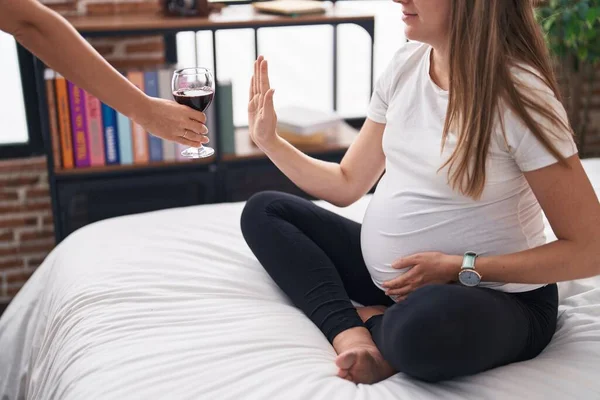 Young pregnant woman doing stop gesture with hand to glass of wine at bedroom