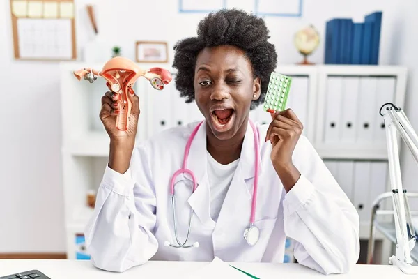 African doctor woman holding anatomical female genital organ and birth control pills winking looking at the camera with sexy expression, cheerful and happy face.