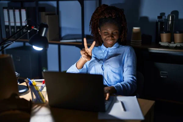 African woman working at the office at night smiling looking to the camera showing fingers doing victory sign. number two.