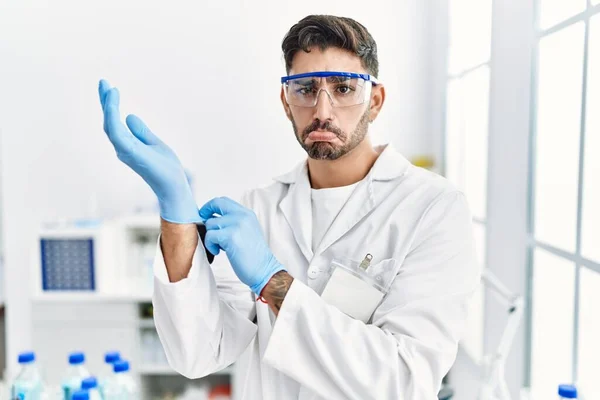 Young hispanic man working at scientist laboratory putting gloves on depressed and worry for distress, crying angry and afraid. sad expression.