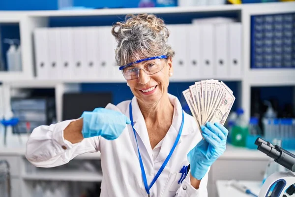 Middle age woman working at scientist laboratory holding dollars pointing finger to one self smiling happy and proud
