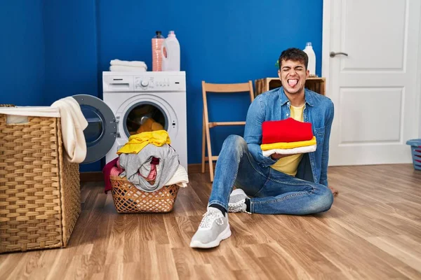 Young Hispanic Man Holding Clean Laundry Sticking Tongue Out Happy — Stock Photo, Image