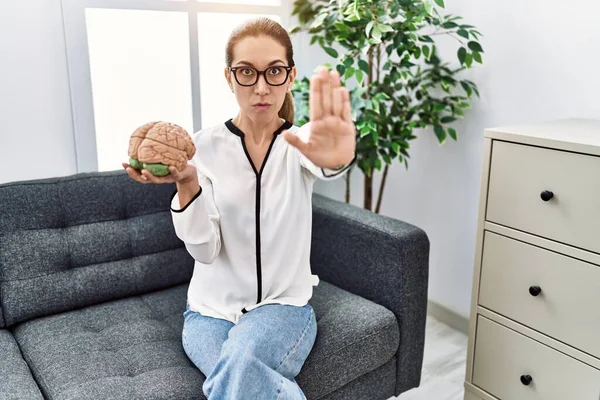 Young hispanic woman holding brain working at psychology clinic with open hand doing stop sign with serious and confident expression, defense gesture