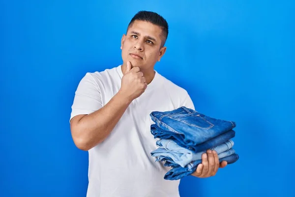 Jeune Homme Hispanique Tenant Pile Jeans Pliés Visage Sérieux Penser — Photo