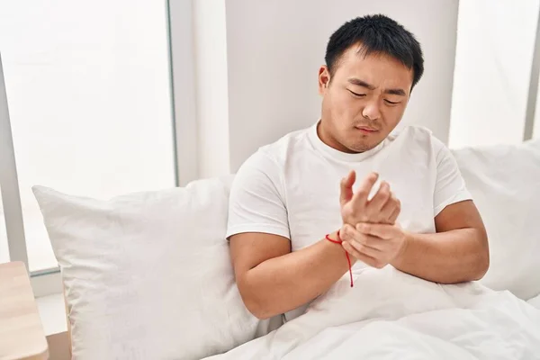 Young chinese man suffering for hand pain sitting on bed at bedroom