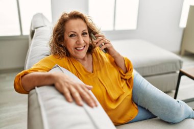 Orta yaşlı beyaz bir kadın akıllı telefonla konuşuyor. Evdeki kanepede oturuyor..