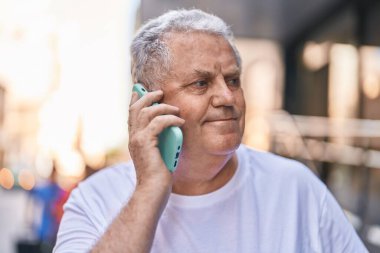 Orta yaşlı, kır saçlı, gülümseyen, kendine güvenen bir adam caddedeki akıllı telefondan konuşuyor.