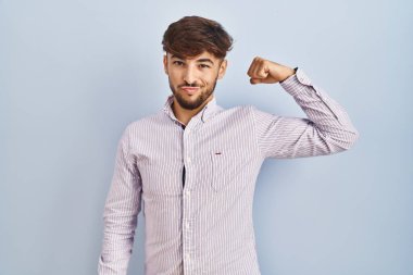 Arab man with beard standing over blue background strong person showing arm muscle, confident and proud of power 