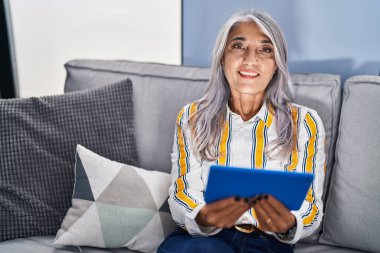 Orta yaşlı, kır saçlı bir kadın dokunmatik yastıkta video izliyor. Evdeki koltukta oturuyor.