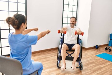 Orta yaşlı bir adam ve kadın fizyoterapi kliniğinde tekerlekli sandalyede oturan dambıllarla rehabilitasyon seansı yapıyorlar.