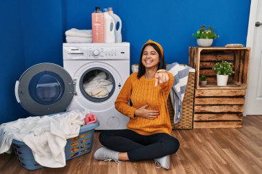 Young hispanic woman doing laundry laughing at you, pointing finger to the camera with hand over body, shame expression 