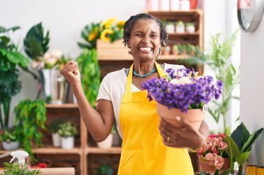 African woman with dreadlocks working at florist shop holding plant screaming proud, celebrating victory and success very excited with raised arm 