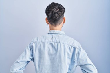 Young hispanic man with beard standing over blue background standing backwards looking away with arms on body 