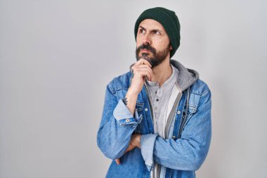 Elleri çenesinde yün şapka takan ve soru soran İspanyol dövmeli genç bir adam, düşünceli bir ifade. Düşünceli bir yüzle gülümsüyor. şüphe kavramı. 