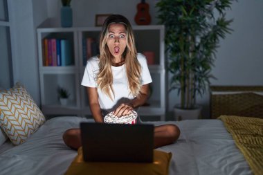 Young blonde woman sitting on the bed at home watching a movie from laptop afraid and shocked with surprise and amazed expression, fear and excited face. 