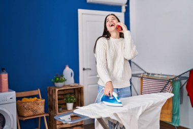 Çamaşırhanede çamaşırları ütüleyen akıllı telefonda konuşan genç bir kadın.