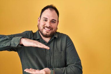 Plus size hispanic man with beard standing over yellow background gesturing with hands showing big and large size sign, measure symbol. smiling looking at the camera. measuring concept. 