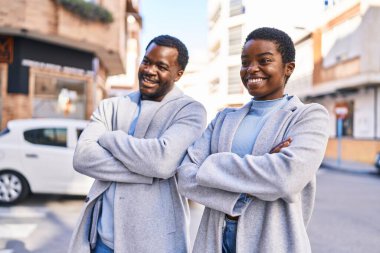 Erkek ve kadın çift gülümsüyor. Kollarını kavuşturup caddede duruyorlar.