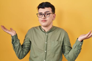 Non binary person standing over yellow background clueless and confused expression with arms and hands raised. doubt concept.  clipart