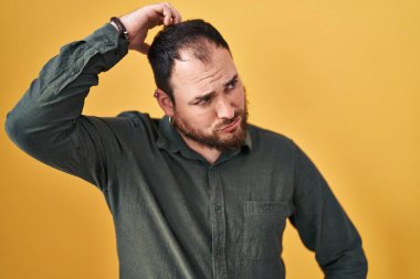 Plus size hispanic man with beard standing over yellow background confuse and wondering about question. uncertain with doubt, thinking with hand on head. pensive concept. 