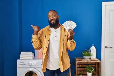 African american man holding money at laundry room pointing thumb up to the side smiling happy with open mouth 