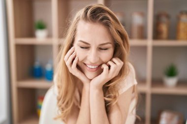 Genç sarışın kadın evde masada kendinden emin bir şekilde gülümsüyor.