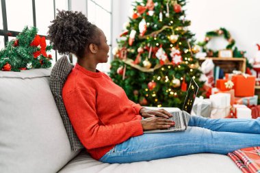Evdeki Noel ağacının yanında dizüstü bilgisayar kullanan Afrikalı Amerikalı kadın.