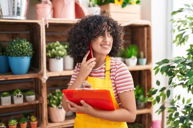 Young middle eastern woman florist talking on smartphone reading clipboard at florist