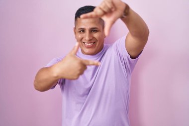 Pembe arka planda duran genç İspanyol adam gülümsüyor ve mutlu yüzlü eller ve parmaklarla çerçeve yapıyor. yaratıcılık ve fotoğrafçılık kavramı. 
