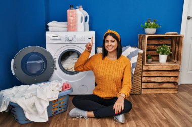 Young hispanic woman doing laundry showing and pointing up with finger number one while smiling confident and happy. 
