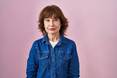 Middle age woman standing over pink background relaxed with serious expression on face. simple and natural looking at the camera. 