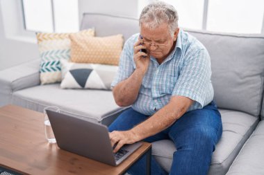 Orta yaşlı, kır saçlı, akıllı telefonlu bir adam evdeki koltukta oturup dizüstü bilgisayar kullanıyor.