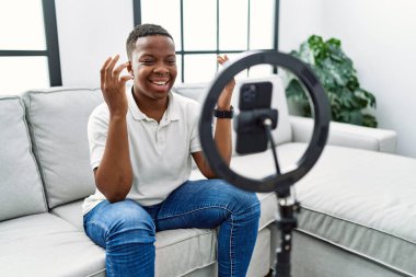 Young african man recording vlog tutorial with smartphone at home celebrating mad and crazy for success with arms raised and closed eyes screaming excited. winner concept 