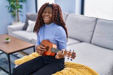 African american woman playing ukulele sitting on sofa at home