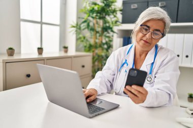 Klinikte doktor üniforması giyen orta yaşlı bir kadın akıllı telefon kullanıyor.