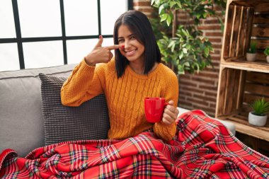Young hispanic woman sitting on the sofa drinking a coffee at home pointing with hand finger to face and nose, smiling cheerful. beauty concept 