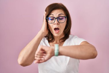 Orta yaşlı İspanyol bir kadın pembe arka planda dikiliyor. Saate bakıyor. Endişeli, geç kalmaktan korkuyor. 