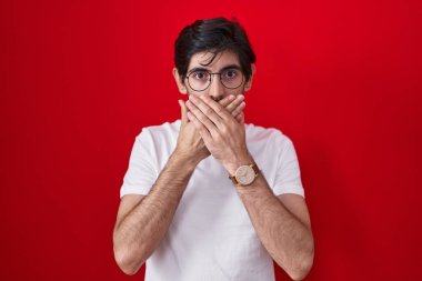 Kırmızı arka planda duran İspanyol genç adam yanlışlıkla ağzını kapatarak şok geçirdi. gizli kavram. 