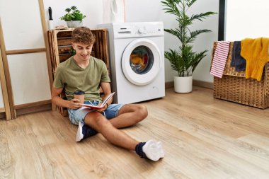 Young caucasian man reading book drinking coffee waiting for washing machine at laundry room
