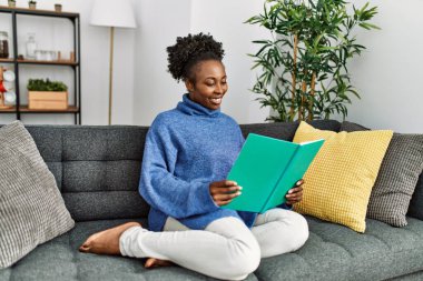 Evdeki kanepede oturan Afro-Amerikalı kadın kitap okuyor.