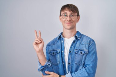 Caucasian blond man standing wearing glasses smiling with happy face winking at the camera doing victory sign with fingers. number two. 