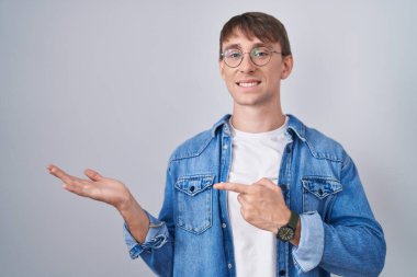 Caucasian blond man standing wearing glasses amazed and smiling to the camera while presenting with hand and pointing with finger. 