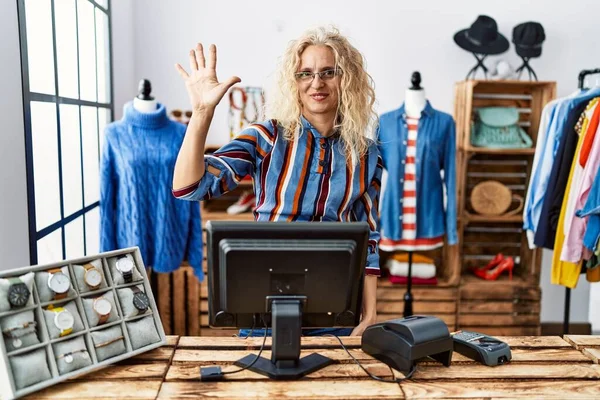 Middle age blonde woman working as manager at retail boutique showing and pointing up with fingers number five while smiling confident and happy.