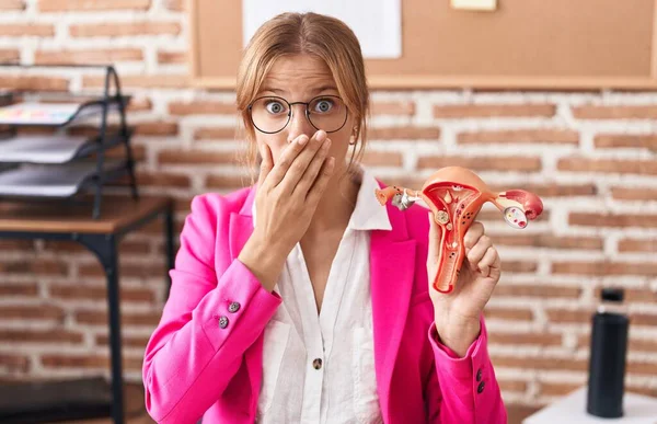 Young caucasian woman holding model of female genital organ at the office complaining for menstruation pain covering mouth with hand, shocked and afraid for mistake. surprised expression