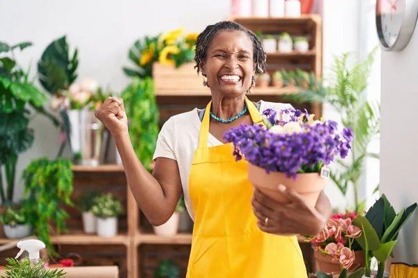 African woman with dreadlocks working at florist shop holding plant screaming proud, celebrating victory and success very excited with raised arm
