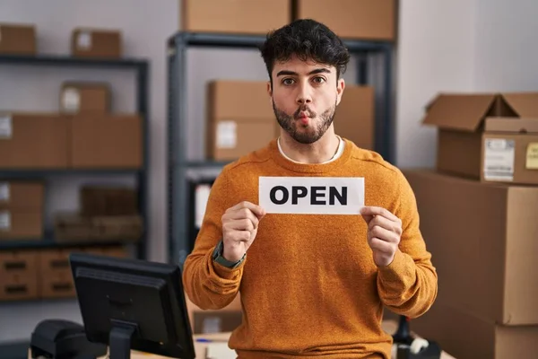 Hispanic man with beard working at small business ecommerce holding open sign making fish face with mouth and squinting eyes, crazy and comical.