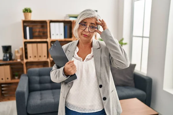 Middle age woman with grey hair at consultation office worried and stressed about a problem with hand on forehead, nervous and anxious for crisis