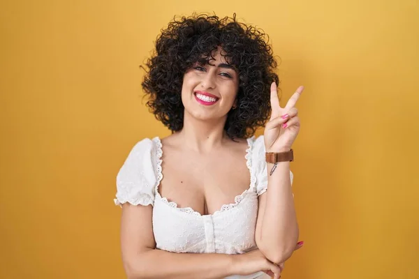 Young brunette woman with curly hair standing over yellow background smiling with happy face winking at the camera doing victory sign with fingers. number two.